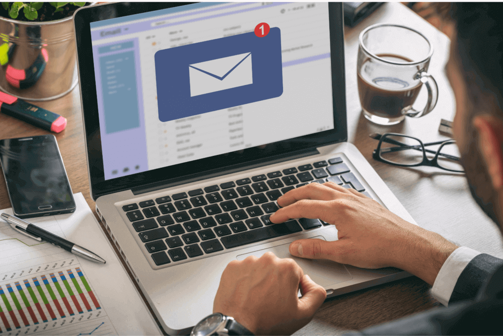 Homme consultant un nouvel email sur son ordinateur portable, en contexte de travail avec graphiques, café et smartphone à proximité.