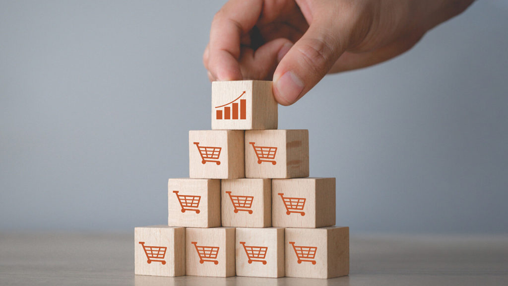 Image représentant une main plaçant un cube avec un graphique de croissance au sommet d’une pyramide de cubes affichant des caddies, illustrant la hiérarchisation et la croissance des sites e-commerce au Maroc.