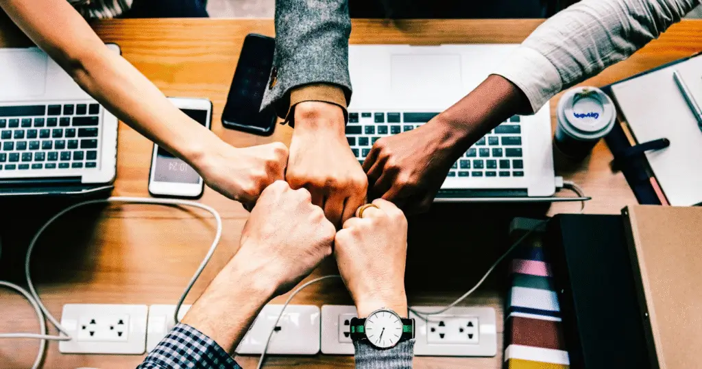 Photo vue de dessus d’une équipe réunie autour d’une table, effectuant un geste de solidarité avec les poings, entourée d’ordinateurs portables, carnets et graphiques, illustrant la collaboration