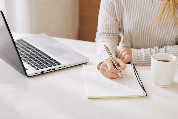 L’image montre une personne assise à une table de travail, prenant des notes dans un carnet ouvert. Un ordinateur portable est allumé à côté d’elle.