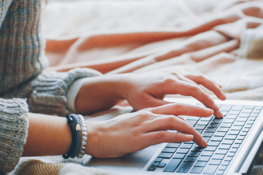 Mains d’une personne tapant sur un clavier d’ordinateur portable, vêtue d’un pull en laine, assise sur un lit recouvert d’une couverture douce.