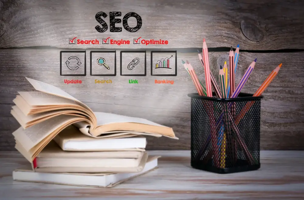 Image illustrant le concept de la formation SEO au Maroc. Elle montre une pile de livres ouverts à gauche, un pot rempli de crayons de couleur à droite, et le mot "SEO" en haut avec les termes "Search", "Engine", "Optimize".